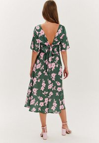 Zelené kvetinové šaty s ružovými kvetmi, V-výstrih, zviazané v páse mašľou. Krátke, ľahké rukávy a dĺžka po polovicu lýtok. Jednoduché ružové sandále.