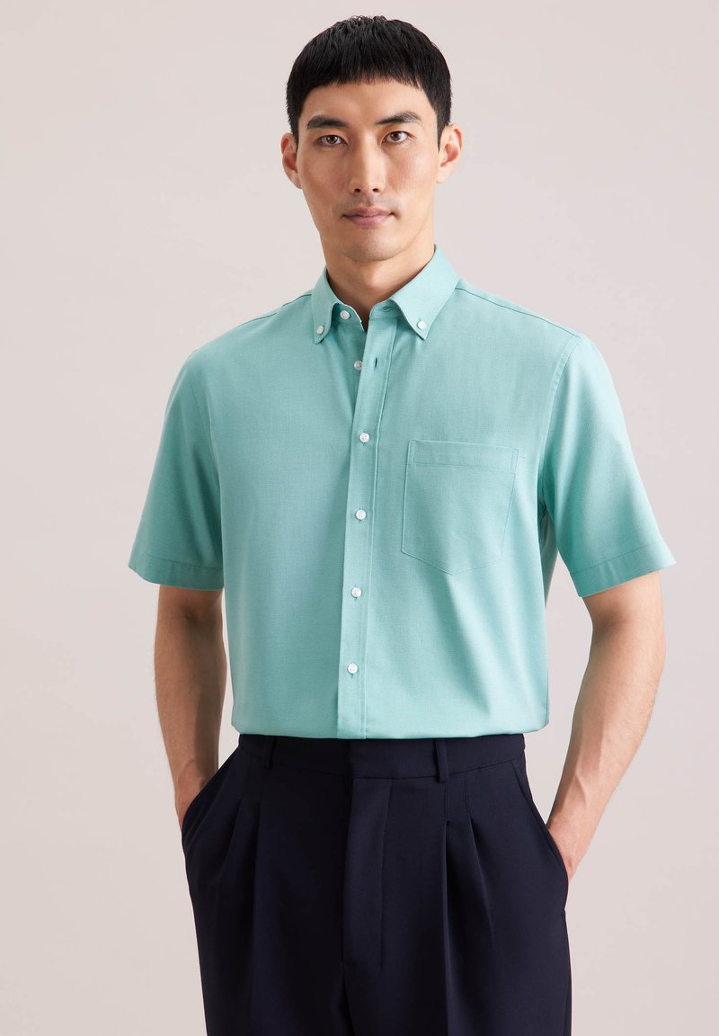 Homme portant une chemise à manches courtes vert menthe avec une poche poitrine et un pantalon plissé bleu marine foncé, debout les mains dans les poches.