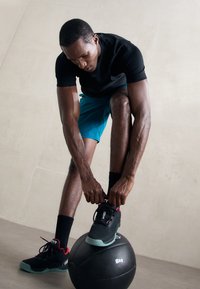 Mężczyzna w czarnej koszuli i niebieskich szortach związujący czarne sneakerki, odpoczywając jedną nogą na 8-kilogramowej piłce do ćwiczeń, przy gładkiej ścianie.