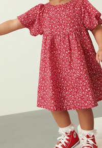 Kind in einem roten Blumenkleid, weißen Rüschenstrümpfen und roten High-Top-Sneakern steht auf einem grauen Boden.