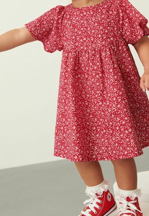 Enfant en robe fleurie rouge, portant des chaussettes blanches avec volants et des baskets montantes rouges, se tenant sur un sol gris.