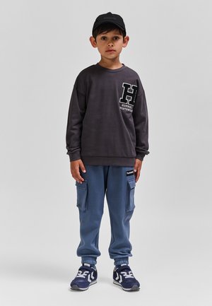 Dunkelgraues Sweatshirt mit weißem Logo, blaue Cargo-Sweatpants mit Taschen und blaue Sneaker mit weißen Akzenten, kombiniert mit einer schwarzen Mütze.