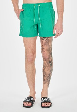 Shorts da bagno verdi con vita elastica e cordino, dotati di tasche laterali. Indossati con ciabatte nere, mostrando un tatuaggio dettagliato sulla gamba.