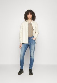 Camicia button-up beige chiaro, canotta marrone, jeans blu e stivali neri. Presenta una texture liscia e una vestibilità casual. Accento in catena d'argento.