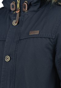 Marineblauwe buitenjas met een gestructureerde stof. Voorzien van een ritssluiting, gouden accenten, leren details en een dubbele knoopsluiting aan de voorkant.