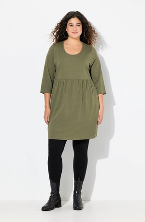 Olivgrünes, knielanges Kleid mit rundem Ausschnitt, Dreiviertelärmeln und gerafftem Taillenbereich. Kombiniert mit schwarzen Leggings und Stiefeletten.