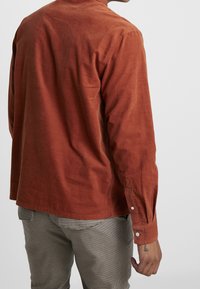 Camisa de pana de color óxido con mangas largas, puños abotonados y un ajuste relajado; combinada con pantalones a rayas texturizados.