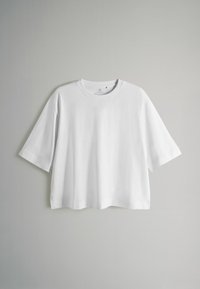 Biała krótko rękawowa koszulka z okrągłym dekoltem, charakteryzująca się luźnym krojem i gładką fakturą, wisząca na szarym tle.