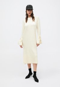 Kremo spalvos megzta suknelė su ribinėmis detalėmis ant rankovių ir hemų; ant riešų yra kaspinėliai. Modelis dėvi juodą kepurę ir batus.