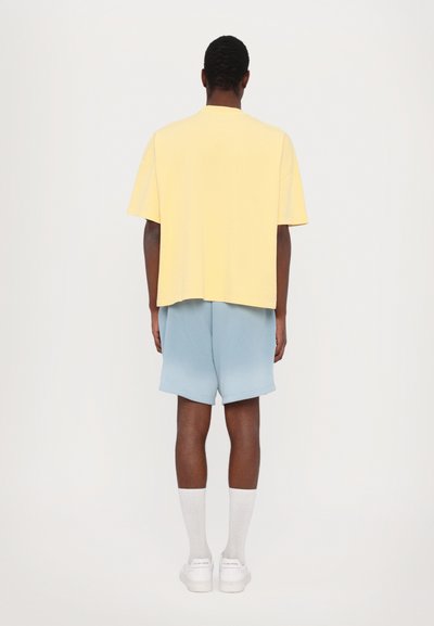 Zachtvallend gele t-shirt, korte mouwen, gecombineerd met lichtblauwe shorts. Het model draagt witte sneakers en kniehoge witte sokken.