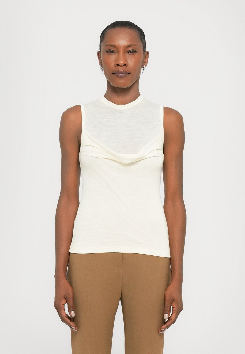 Sleeveless cream top with a high neckline, draped front detail, and smooth texture. Paired with fitted brown pants.