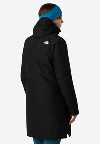 Čierna izolovaná parka s kapucňou, s logom na ľavom pleci a rovnom strihom. Nosená s modrým čelenkou a tyrkysovými nohavicami.
