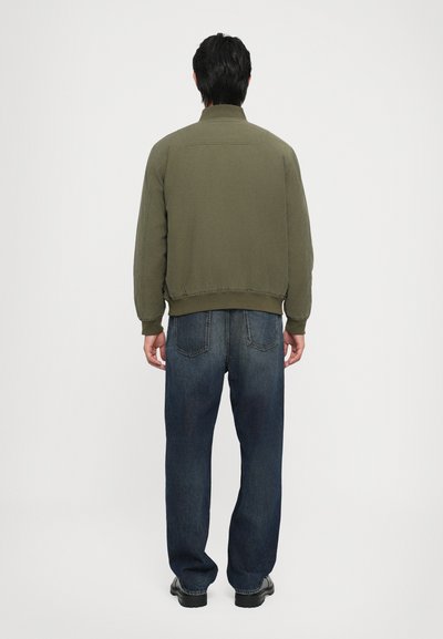 Olivgrön jacka med avslappnad passform och ribbade ärmslut, kombinerad med mörka denimjeans med bakfickor. Enkelt, minimalistiskt design.