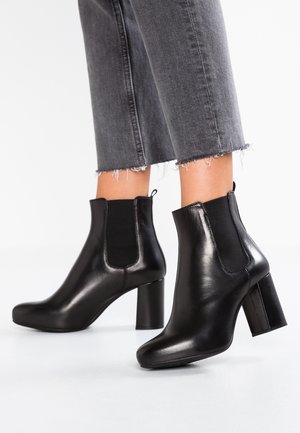 Sorte ankelstøvler i læder med blokhæle båret sammen med grå jeans med frayed kant og kort ben på hvid baggrund.