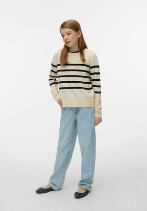 Młoda osoba stojąca, ubrana w kremowy sweter z czarnymi poziomymi paskami, jasnoniebieskie dżinsy oraz czarne baleriny z wzorem na białym tle.