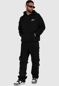 Zwarte hoodie met een voorzak en logo, gecombineerd met zwarte cargo pants met zijzakken. G gedragen met zwart-witte sneakers.