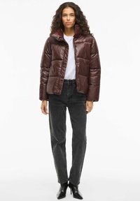 Smeđi puffer kaput s sjajnim završetkom, visokim ovratnikom i zatvaranjem na patentni zatvarač, uparen s crnim trapericama visokog struka i crnim čizmama s oštrom kapicom.