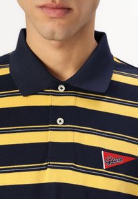 Marineblå og gulstribet polo shirt med krave, der har et lille broderet rødt logo og to knapper ved halsen. Struktureret stof.
