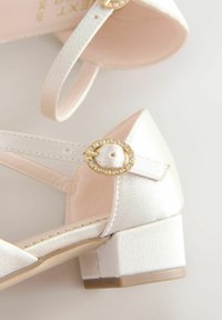 Witte satijnen schoenen met lage hakken en enkelbandjes, gekenmerkt door ovale gouden gespen versierd met heldere rhinestones.