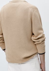 Beżowy sweter wykonany na drutach z teksturowanej tkaniny. Posiada opadające szwy ramion oraz podwinięte mankiety z czarnym akcentem na dole.