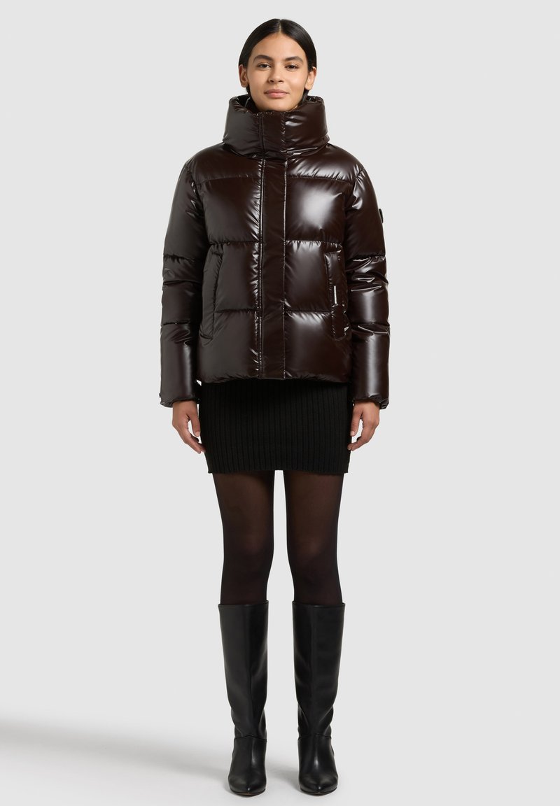 Brown puffer jacket with a high collar, glossy finish, stitched pattern, and side pockets, worn over a black ribbed skirt and knee-high boots.