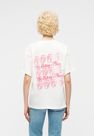 Persona con capelli corti, ricci e biondi che indossa una maglietta bianca con loghi rosa dei Rolling Stones e testo sul retro, abbinata a jeans azzurri.