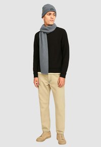Uomo che indossa un berretto grigio e una sciarpa, un maglione nero, pantaloni beige e scarpe beige, in piedi contro uno sfondo grigio chiaro uniforme.
