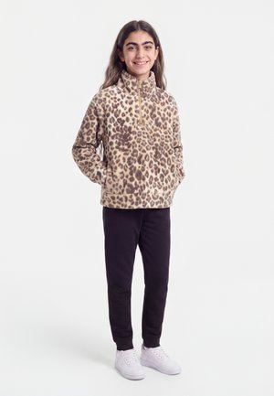 Jeune personne souriante, portant une polaire zippée imprimé léopard, pantalon noir et baskets blanches, debout les mains dans les poches sur fond blanc.