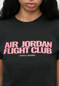 Μαύρο μπλουζάκι με ροζ κείμενο "AIR JORDAN FLIGHT CLUB" και μικρότερο λευκό κείμενο "OFFICIAL MEMBER" που φοράει ένα άτομο με μακριά σκούρα μαλλιά.