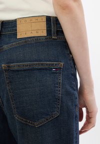 Sötétkék farmernadrág, világosbarna bőrpacsni a hátsó derékpánton, rajta "TOMMY JEANS" felirattal és egy kis zászló részlettel a zsebében.