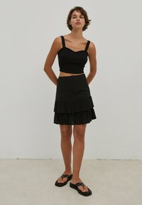 Zwarte geribbelde crop top met brede banden, gecombineerd met een zwarte gelaagde rokken met ruches. Model draagt zwarte slippers en staat tegen een neutrale achtergrond.