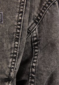 Jeansjacka i svart med vit söm, med en närbild av texturerat tyg, synliga sömmar och lätt slitna kanter.