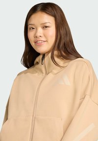 Beige rits-hoodie met een hoge kraag, gemaakt van gemêleerd stof. Heeft grote voorzakken en drie lichtgekleurde strepen op de borst.