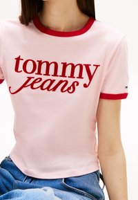 Rosa kortärmad skjorta med röd ribbad halsringning och manschetter. "tommy jeans"-logotyp i fet röd text över framtill. Mjuk bomullsmaterial.