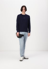 Chemise à manches longues marine avec le logo "GAP", jeans bleu clair, pochette noire à la taille, et baskets blanches avec des accents gris. Fond neutre.