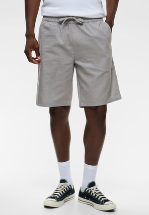 Man die een grijze short met trekkoord, witte sokken en zwarte lage sneakers draagt, staat met handen in de zakken.