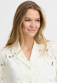 Vit spetsblus med blommönster, knappstängning och krage. Mjuk textur och genomskinlig design.