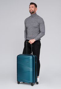 Maleta rígida color teal con líneas diagonales texturizadas, mango retráctil y cuatro ruedas. Diseñada para viajar y facilitar el movimiento.