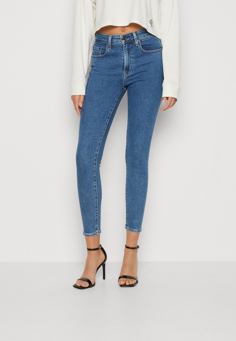 Jean skinny en denim bleu taille haute, avec cinq poches et une coupe ajustée. Porté avec des sandales à talons noires et un top blanc court.