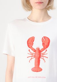 Camiseta blanca que presenta un gráfico audaz de una langosta roja y el texto "SÓLO LLÁMAME EGÓISTA" debajo, fabricada con una suave tela de algodón.