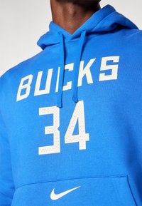 Mėlyna džemperis su kapuce, ant kurio užrašas "BUCKS" ir skaičius "34" balta spalva. Džemperis turi raištelius kapucei ir Nike logotipą ant priekinės kišenės. Sklandi medžiagos tekstūra.