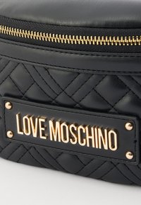 Črna prešita usnjena torba z zlatim zadrgo in vidno zlato ploščico z napisom "LOVE MOSCHINO." Struktura površine.