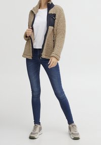 Personne portant une veste polaire beige, un t-shirt blanc, un jean slim bleu et des baskets beiges, debout devant un fond uni.