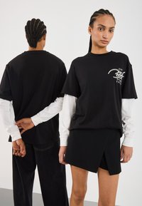 Sort oversized t-shirt med grafisk tryk foran og lagte hvide lange ærmer, parret med en sort omslagsskjørt. Model poserer fra bag og foran.