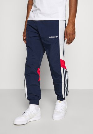 Mężczyzna ubrany w granatowe spodnie dresowe Adidas z białymi i czerwonymi paskami, w zestawie z białymi butami sportowymi Adidas i białą koszulką.