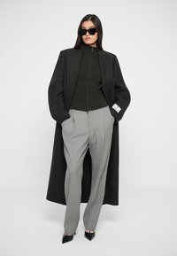 Casaco preto longo sobre uma blusa verde-escura, combinado com calças cinza claro de corte elegante e sapatos de salto agudo pretos. Acessórios incluem óculos de sol escuros.