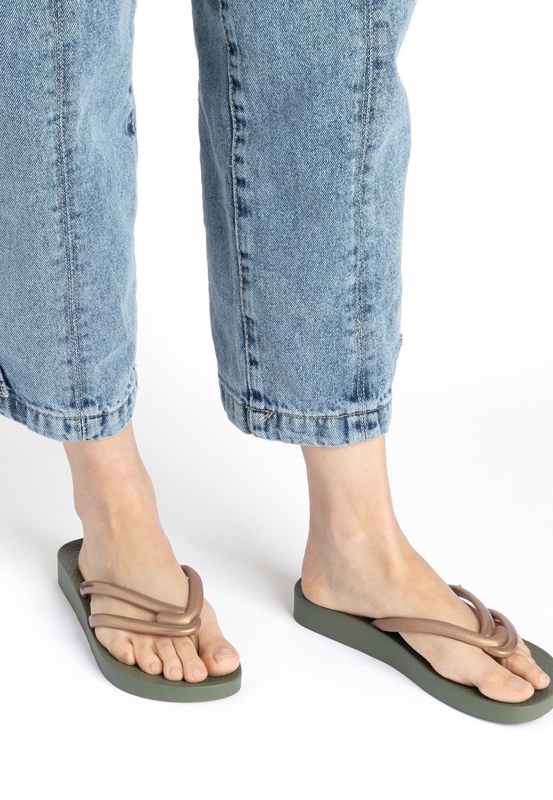 Flip-Flops mit einer grünen Gummisohle und einem braunen, geschlungenen, strukturierten Riemen. Getragen mit hellblauen, verkürzten Jeanshosen.