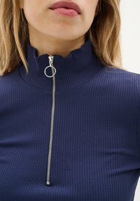 Top bleumarin cu dungi, cu guler înalt și închidere cu fermoar argintiu, având un tab de tip circular. Țesătura texturată adaugă profunzime designului.