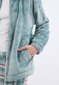 Ręka w kieszeni miękkiej, jasnoniebieskiej polarowej kurtki noszonej na białej prążkowanej bluzce i zielonych spodniaczkach w kratę na jednolitym białym tle.