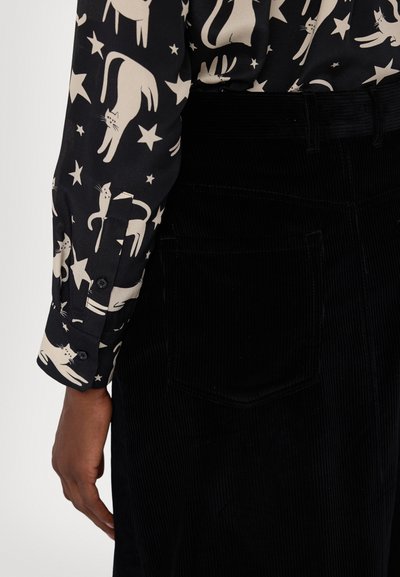 Pantaloni in velluto a coste neri abbinati a una camicia a maniche lunghe con un motivo di gatti color crema e stelle. La camicia ha polsini con bottoni.